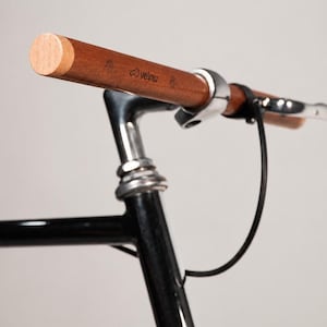 Pode incluir: Um guiador de bicicleta feito de madeira castanha com uma tampa de extremidade de cor clara, apresentando a palavra "velow" impressa na superfície. O guiador está fixado a uma estrutura de metal preto com uma campainha de metal prateado.