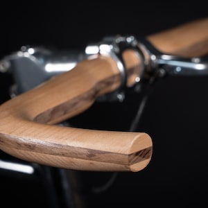 Pode incluir: Um guiador de bicicleta feito de madeira de tom claro com um padrão de grão visível. O guiador apresenta um design curvo e está fixado ao quadro da bicicleta com braçadeiras e ferragens de metal prateado polido sobre fundo escuro.