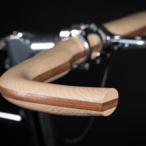 Puede incluir: Un manillar de bicicleta fabricado en madera clara con una incrustación central de madera oscura, que presenta un diseño curvo y suave. El manillar está fijado de forma segura a una potencia de metal plateado, mostrando la veta natural.