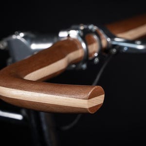 Op de afbeelding: Een fietsstuur gemaakt van gelaagd donker en licht hout, met een gebogen ontwerp en een gladde afwerking. Het houten stuur is bevestigd aan een metalen zilverkleurige stuurpen, wat het contrast tussen de houtnerf en het metaal benadrukt.