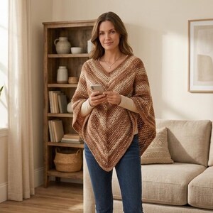 Puede incluir: Un poncho de ganchillo marrón y crema con cuello en V. El poncho tiene un diseño a rayas y se lleva sobre una camisa crema de manga larga. La persona lleva vaqueros azules y sostiene un smartphone.