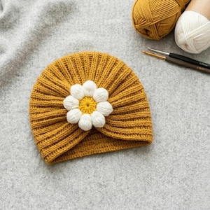 May include: Mustard yellow knitted turban-style hat with a white and yellow daisy applique. The hat is made of a ribbed knit fabric. Yarn and crochet hooks are in the background.