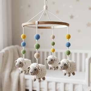 May include: A handmade baby mobile featuring four crocheted sheep suspended below a wooden ring. Colorful crochet balls in yellow, blue, green, and beige are strung above each sheep. The mobile hangs in front of a white crib.