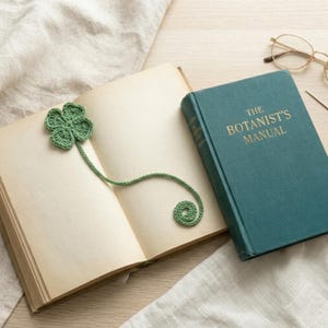 Könnte beinhalten: Ein grünes, gehäkeltes vierblättriges Kleeblatt-Lesezeichen liegt in einem geöffneten Buch. Das Buch hat gealterte Seiten und liegt neben einem teal-farbenen Buch mit dem Titel "The Botanist's Manual". Goldgerahmte Brille und ein Stift liegen auf der Holzoberfläche.