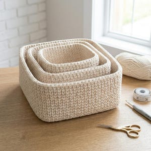 May include: A set of three cream-colored, square-shaped crochet baskets nested together. The baskets are made of a thick, textured yarn. A ball of yarn, scissors, a crochet hook, and a measuring tape are on the wooden surface.
