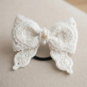 Peut inclure: Un chouchou blanc en crochet en forme de nœud papillon, orné de perles et d'une fleur dorée. Le nœud présente des détails de dentelle complexes et est fixé à un élastique noir. Cet accessoire fait main ajoute une touche d'élégance.
