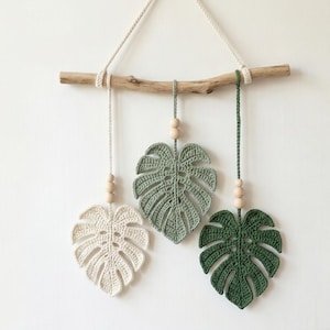 May include: Three crocheted Monstera leaf wall hangings in cream, sage green, and dark green, suspended from a driftwood branch with wooden beads. The leaves are intricately detailed, showcasing a tropical theme. The wall decor is hung with natural twine.