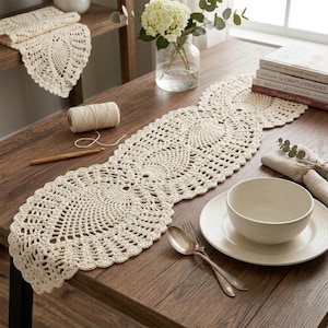 May include: Cream-colored, crocheted table runner with a scalloped edge, displayed on a wooden table. The runner features a repeating circular pattern. A matching doily is on a shelf in the background. A bowl, plate, and silverware are on the table.