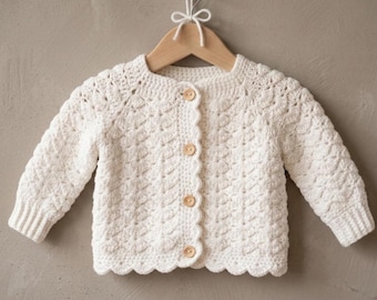 May include: A cream-colored, crocheted baby cardigan with long sleeves and a scalloped edge. The sweater has a textured pattern and is fastened with four wooden buttons. It hangs on a wooden hanger.