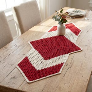 Puede incluir: Camino de mesa de ganchillo rojo y crema hecho a mano. El camino de mesa presenta un diseño geométrico con una forma de diamante rojo central y bordes crema. Un pequeño jarrón blanco con vegetación y bayas se encuentra sobre el camino de mesa, sobre una mesa de madera.