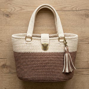 May include: A two-tone crochet tote bag in cream and brown. The bag features a rectangular shape with a flap closure and a decorative tassel. The handles are cream-colored and attached with gold-colored rings.