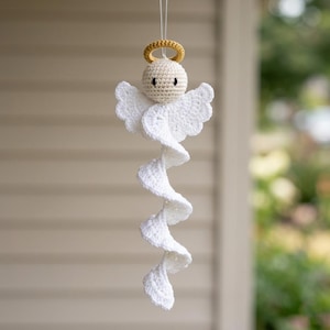 May include: A hanging crochet angel ornament featuring a round beige head with black eyes, a yellow halo, and white wings. The body consists of a long, white, spiraling crochet tail that hangs downward, suspended by a thin, clear filament string.