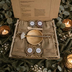 May include: A skincare set containing natural clay powders for face masks. The set includes small burlap bags of clay, small jars of powder, a wooden bowl, a brush, and a wooden spoon. The box is open, revealing the contents. The text on the box reads "Natural clay skincare powders for face mask applications."