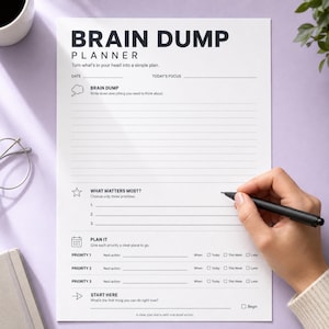 May include: A white Brain Dump Planner sheet with black text and lines. The page includes sections for Date, Today's Focus, Brain Dump, What Matters Most, Plan It, and Start Here. A black pen is writing on the paper, which features checkboxes.