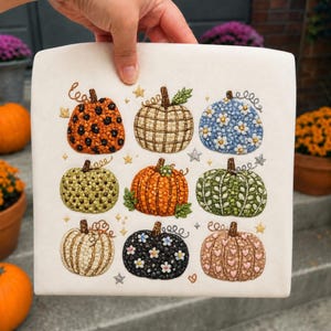 May include: A white fabric square featuring nine embroidered pumpkins in a grid. The pumpkins display various patterns including leopard print, plaid, floral, polka dots, and hearts in colors like orange, green, blue, black, and pink, accented by small stars.
