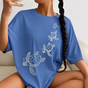 Puede incluir: Una camiseta azul de manga corta con un estampado gráfico blanco de cuatro tortugas marinas de diferentes tamaños nadando en un patrón diagonal ascendente. Las tortugas muestran patrones detallados en sus caparazones y pequeñas burbujas.