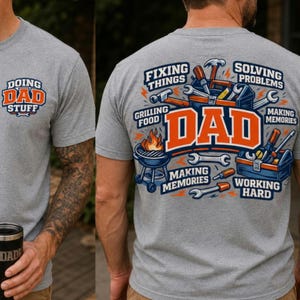 May include: A heather gray t-shirt featuring a small logo on the front that reads "DOING DAD STUFF". The back displays a large graphic with the word "DAD" in orange, surrounded by illustrations of tools, a grill, and text including "FIXING THINGS", "SOLVING PROBLEMS", "GRILLING FOOD", "MAKING MEMORIES", and "WORKING HARD".