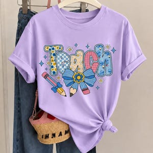 Pode incluir: Uma t-shirt roxo-claro com a palavra "Teach" em letras grandes com padrões florais, xadrez e tartã. Abaixo do texto, há um laço azul e dois lápis. A t-shirt está atada na cintura e é apresentada com calças de ganga azuis.