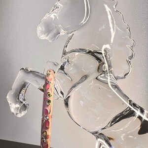 Puede incluir: Una escultura de caballo de cristal transparente con un bolígrafo rosa y dorado. El bolígrafo está decorado con diseños florales rosas y pequeños detalles plateados y dorados. El caballo está encabritado.