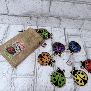 Puede incluir: Una bolsa de arpillera beige con cordón y el texto Ladybug Ladybug impreso en el frente, acompañada de seis fichas de mariquita de madera en verde, morado, azul, naranja, rojo y amarillo, además de un pequeño dado blanco de seis caras con puntos negros.