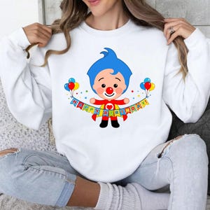May include: A white long-sleeve sweatshirt featuring a graphic of a cartoon clown with blue hair and a red nose. The clown wears a red cape and holds a colorful banner that reads "HAPPY BIRTHDAY!" with balloons on each side of the character.