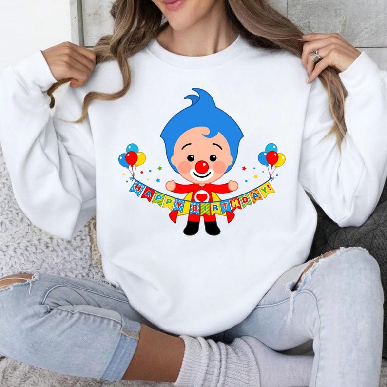 May include: A white long-sleeve sweatshirt featuring a graphic of a cartoon clown with blue hair and a red nose. The clown wears a red cape and holds a colorful banner that reads "HAPPY BIRTHDAY!" with balloons on each side of the character.