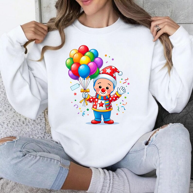 May include: A white long-sleeve sweatshirt featuring a graphic of a cartoon clown wearing a red and white polka dot shirt, a blue scarf, and a matching hat. The clown holds a large bunch of colorful balloons, surrounded by small confetti shapes.