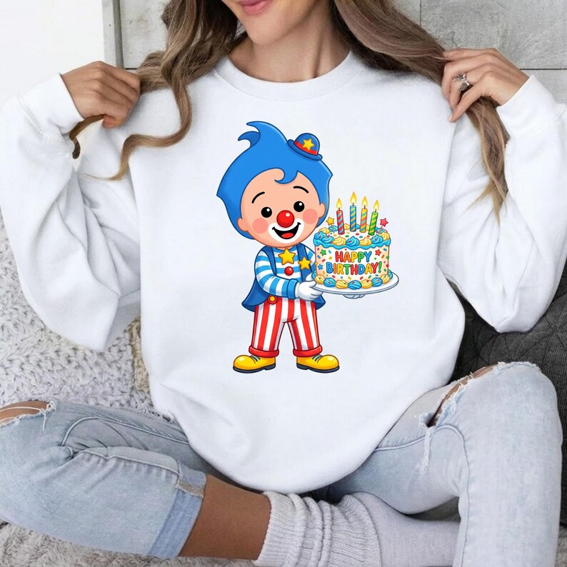 May include: A white long-sleeve sweatshirt featuring a graphic of a cartoon clown with blue hair, a red nose, and a blue and white striped shirt. The clown holds a birthday cake with lit candles and the text Happy Birthday printed on the front.