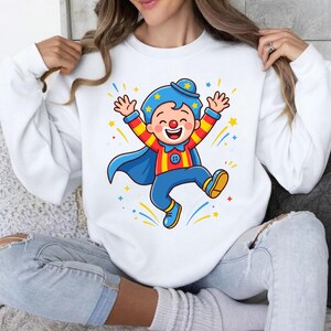May include: A white long-sleeve sweatshirt featuring a colorful graphic of a cartoon clown wearing a blue hat, a red and yellow striped shirt, blue pants, and a blue cape, with yellow stars and blue and yellow streaks surrounding the figure.