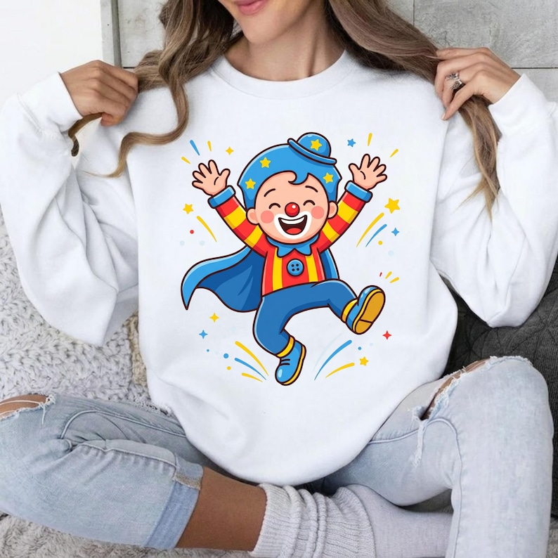 May include: A white long-sleeve sweatshirt featuring a colorful graphic of a cartoon clown wearing a blue hat, a red and yellow striped shirt, blue pants, and a blue cape, with yellow stars and blue and yellow streaks surrounding the figure.