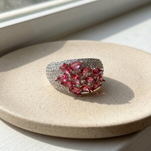 May include: A wide silver-toned ring featuring a cluster of pear-shaped pink gemstones arranged in a floral pattern, surrounded by numerous small, clear pav&eacute;-set stones that cover the remaining surface of the band for a sparkling finish.