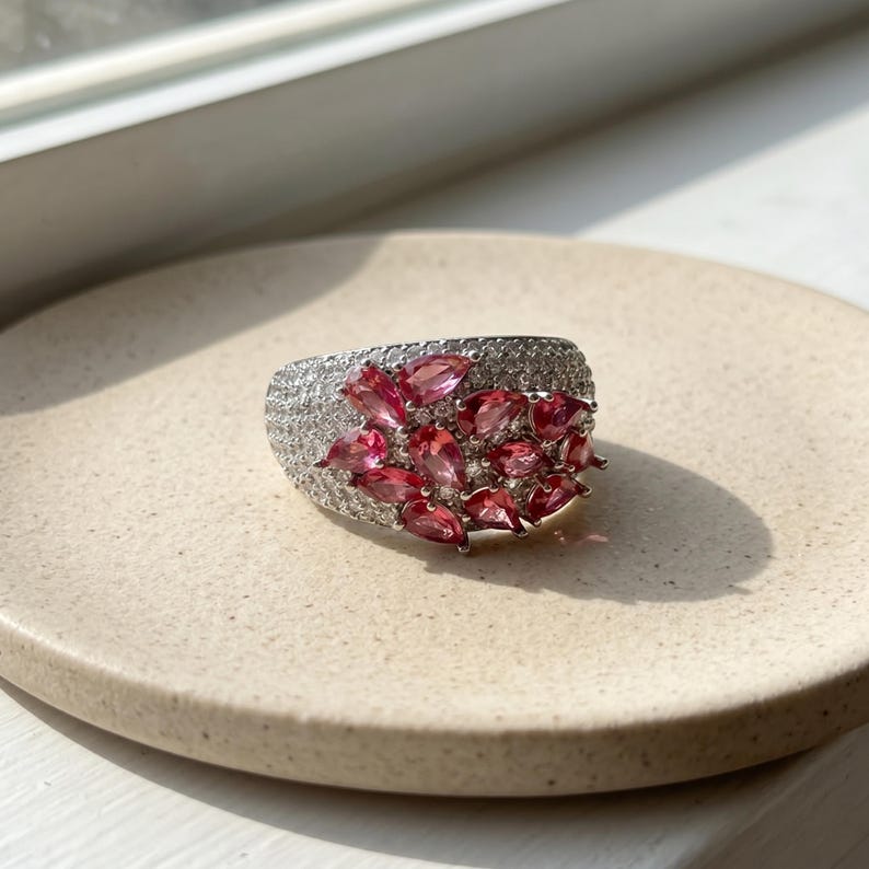 May include: A wide silver-toned ring featuring a cluster of pear-shaped pink gemstones arranged in a floral pattern, surrounded by numerous small, clear pav&eacute;-set stones that cover the remaining surface of the band for a sparkling finish.
