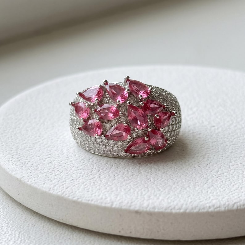 May include: A wide silver ring featuring a cluster of pear-shaped pink gemstones set against a background of numerous small, clear pav&eacute;-set stones that cover the entire surface of the band, creating a dense and sparkling appearance.