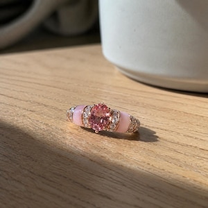 May include: A rose gold ring with a central oval-cut pink gemstone, flanked by smaller clear stones and pink enamel accents. The ring is set on a wooden surface, with a white mug in the background.
