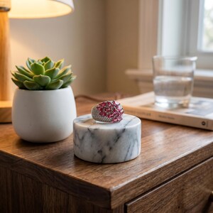 May include: A silver-toned ring featuring a cluster of marquise-cut pink stones and small clear pav&eacute; stones, resting on a cylindrical white marble display stand with grey veining, showcasing a detailed and intricate design on the band.