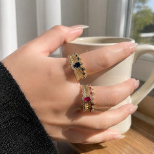 Puede incluir: Dos anillos dorados con forma de corona y piedras preciosas de colores. Un anillo presenta una gran piedra azul ovalada, el otro una piedra roja. Ambos anillos tienen piedras de acento más pequeñas y se muestran en una mano.