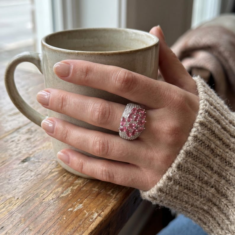 May include: A wide silver ring featuring a cluster of marquise-cut pink gemstones surrounded by small clear pav&eacute; stones. The ring has a textured, metallic band that wraps around the finger, showcasing a prominent, multi-faceted pink stone arrangement.
