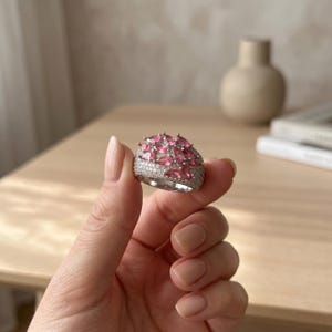 May include: A silver-toned ring featuring a wide band encrusted with numerous small clear stones. The top of the ring is adorned with a cluster of multiple pear-shaped pink stones arranged in a dense, layered formation across the surface.