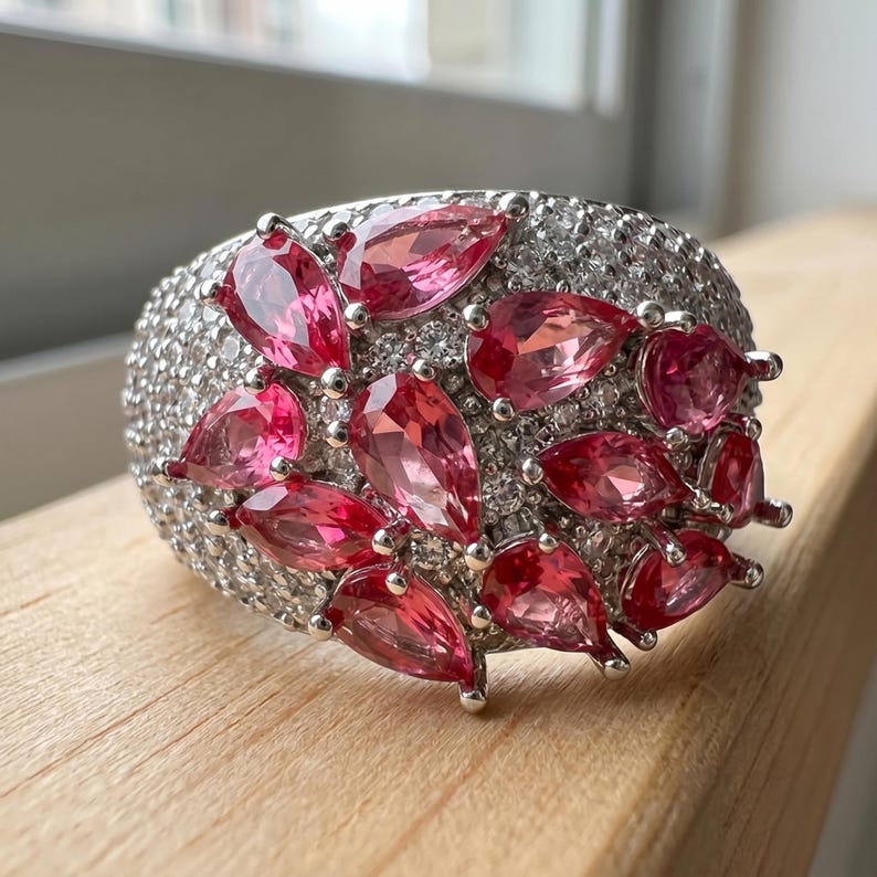May include: A wide silver-toned ring featuring a cluster of pear-cut pink gemstones set against a background of numerous small, clear, round-cut stones that cover the entire surface of the band, creating a dense, sparkling, and textured appearance.