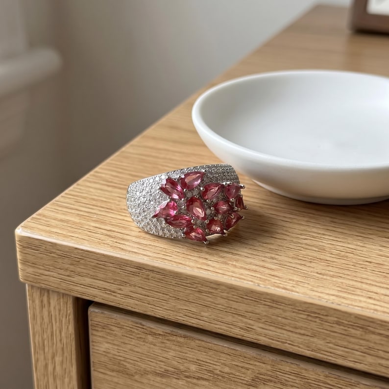 May include: A wide silver-toned ring featuring a cluster of pear-shaped pink gemstones arranged in a floral pattern, surrounded by a pav&eacute; setting of numerous small, clear, sparkling stones that cover the entire surface of the band.