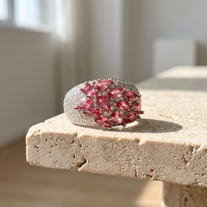 May include: A silver-toned ring featuring a cluster of pear-shaped pink gemstones arranged in a floral pattern, surrounded by numerous small, clear pavé-set stones covering the wide band surface, creating a textured and reflective appearance.