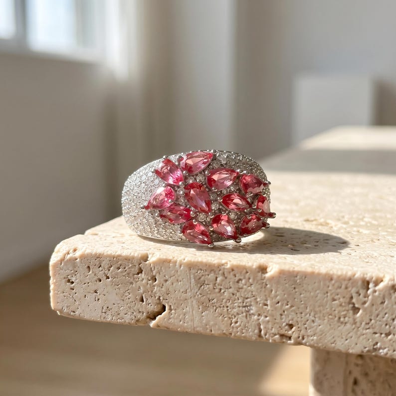 May include: A silver-toned ring featuring a cluster of pear-shaped pink gemstones arranged in a floral pattern, surrounded by numerous small, clear pav&eacute;-set stones covering the wide band surface, creating a textured and reflective appearance.