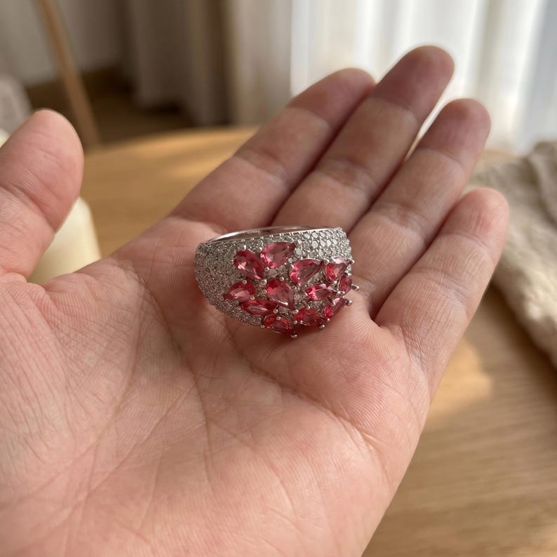 May include: A wide silver-toned ring featuring a cluster of pear-shaped pink gemstones set against a background of numerous small, clear pav&eacute;-set stones that cover the entire surface of the band, creating a dense and sparkling appearance.