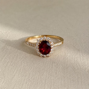 May include: A gold ring featuring a large, oval-cut, deep red gemstone surrounded by a halo of smaller, clear gemstones. The band is also adorned with smaller gemstones. The ring is set against a neutral, textured background.