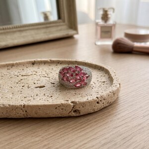 May include: A silver-toned ring featuring a cluster of pear-shaped pink gemstones surrounded by small, clear pav&eacute;-set stones. The ring has a wide, rounded band design that covers the top of the finger with a dense arrangement of pink and clear stones.