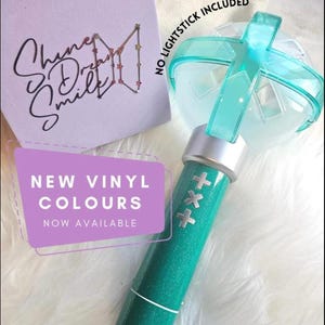 Può includere: Un manico per lightstick color verde acqua glitterato con un colletto argentato e una parte superiore traslucida a forma di croce. Un cartoncino quadrato viola riporta il testo Shine Dream Smile e un logo a stelle. Il testo recita NO LIGHTSTICK INCLUDED e NEW VINYL COLOURS NOW AVAILABLE.