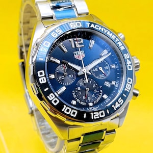 May include: A stainless steel TAG Heuer Formula 1 watch with a blue dial and bezel. The watch features a chronograph function and a tachymeter scale. The watch band is also stainless steel.