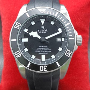 Könnte beinhalten: Eine Tudor Pelagos Armbanduhr mit schwarzem Zifferblatt, weißen lumineszierenden Stundenmarkierungen und einer schwarzen drehbaren Lünette. Das Zifferblatt zeigt den Text Tudor Geneve, Pelagos, Chronometer Officially Certified, Rotor Self-Winding, 500m-1640ft und Swiss Made. Sie hat ein schwarzes Kautschukarmband.
