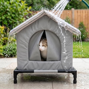 May include: A gray, A-frame pet shelter with a peaked roof, supported by a black tubular frame. A tabby cat is visible inside the arched entrance. Water is being sprayed over the top of the structure, creating a cascading effect down the sides.