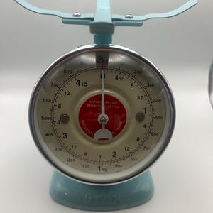 May include: A vintage-style, light blue kitchen scale with a round face and a curved handle. The scale features measurements in pounds and ounces, with a maximum capacity of 4 pounds. The face has a clear glass cover and a red center.